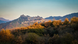 Borgo medievale arroccato su una collina rocciosa con paesaggio autunnale e cielo azzurro