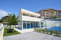 Struttura sportiva bianca con grande vetrata, terrazza, giardino con alberi e campo da padel
